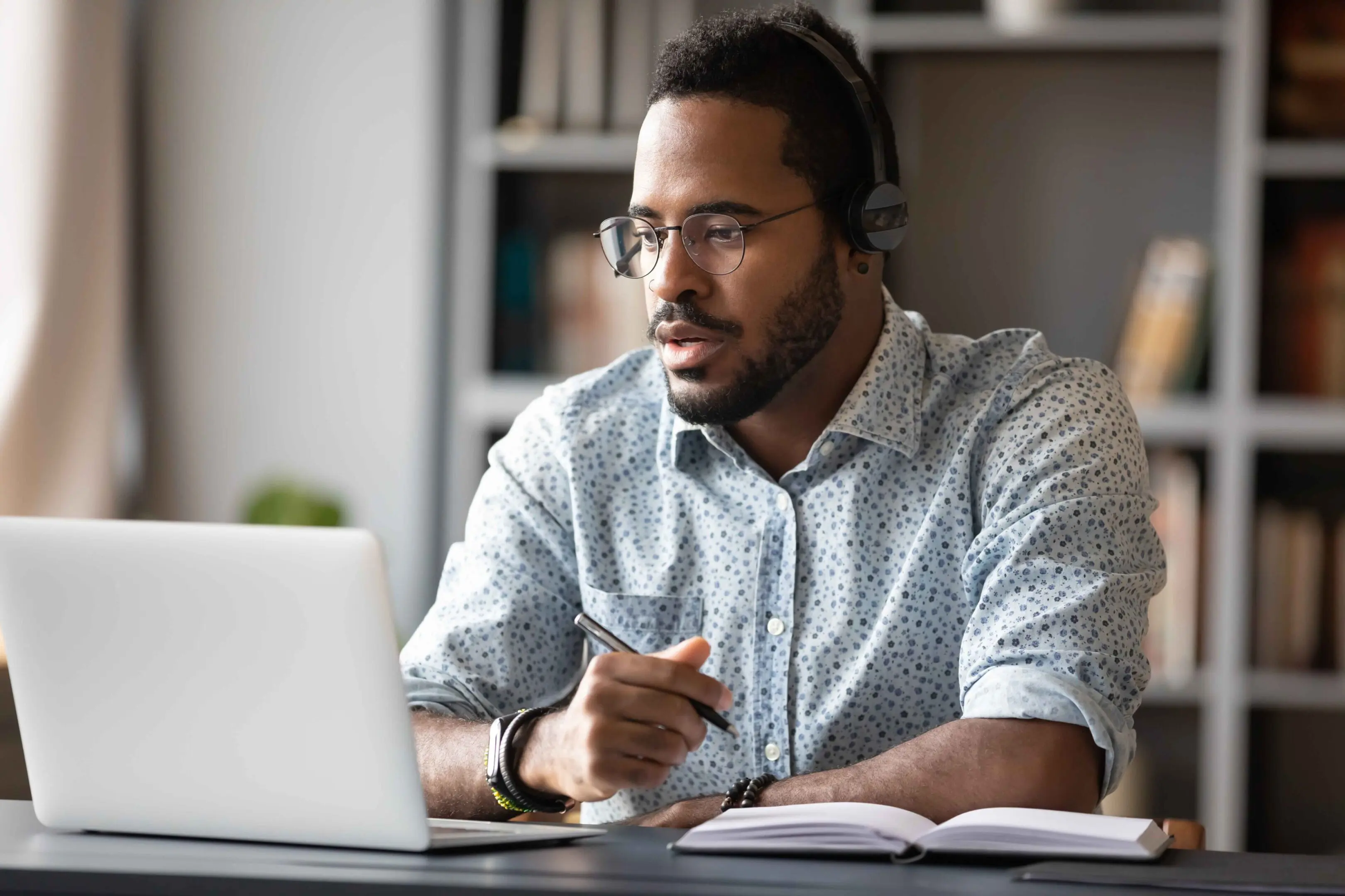 GettyImages-1203044233_11zon Man wearing headphones and glasses sits at a desk, looking at a laptop, holding a pen, with an open notebook in front of him, a bookshelf in the background.