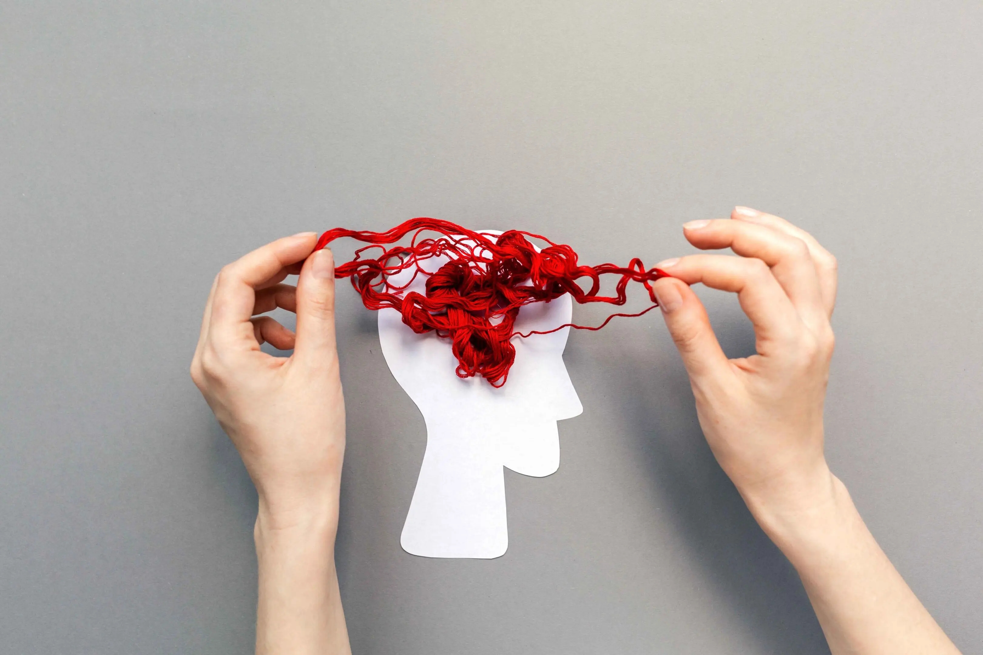 GettyImages-1338451012_11zon Two hands hold tangled red yarn above a white paper cutout of a human head, against a plain gray background.