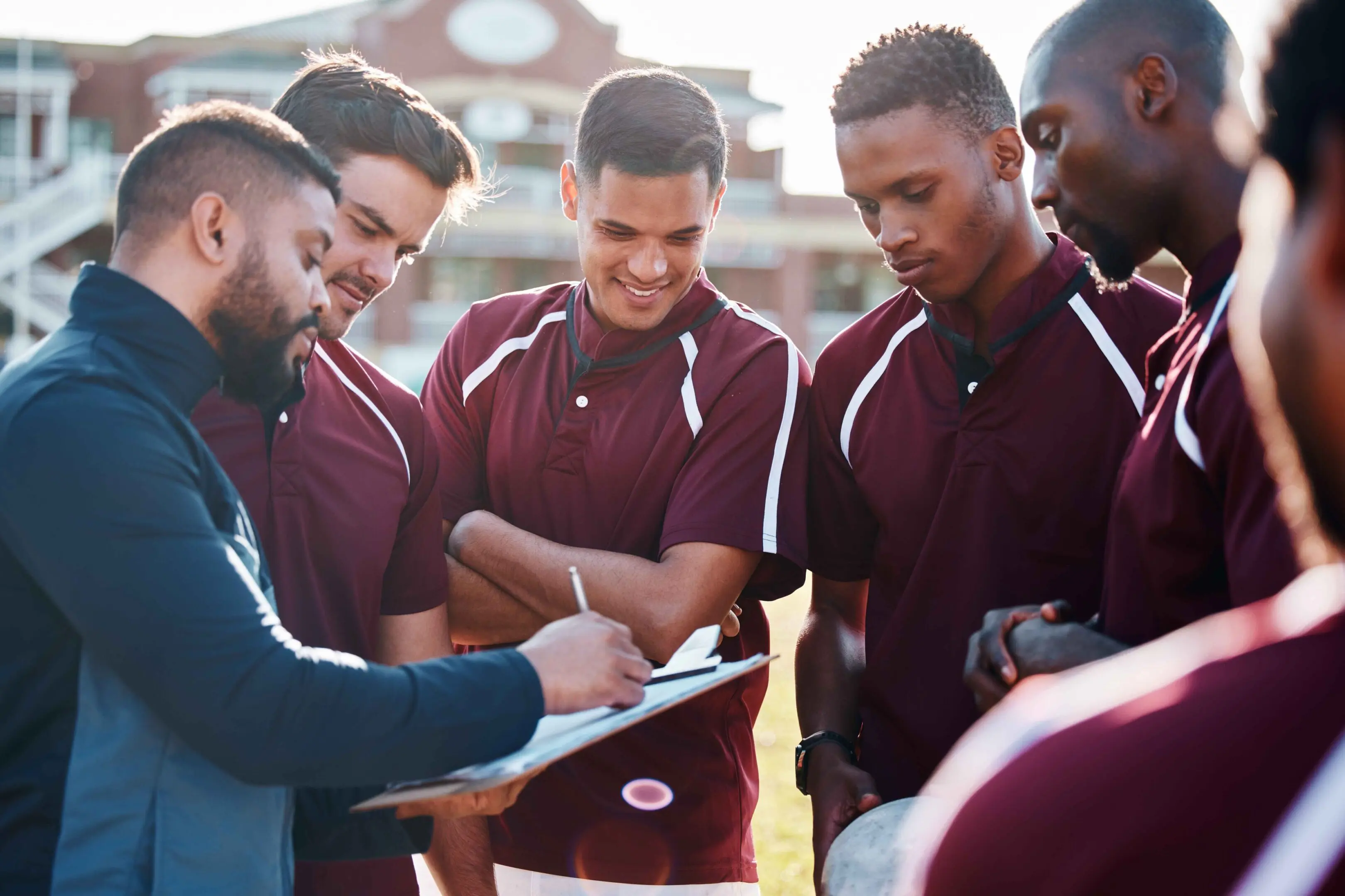 A coach discusses strategy with a group of male athletes wearing maroon uniforms outdoors, using a clipboard during a team meeting.