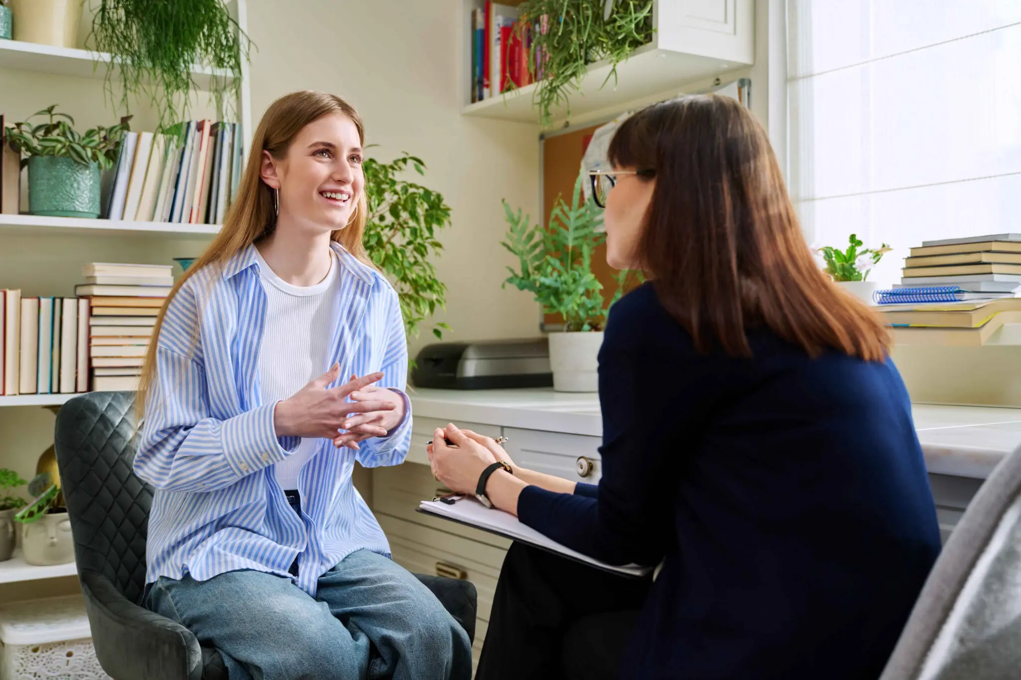 GettyImages-2148493228_11zon Two women sit facing each other in a well-lit office, engaged in conversation. One is smiling and talking, while the other listens and takes notes on a clipboard.