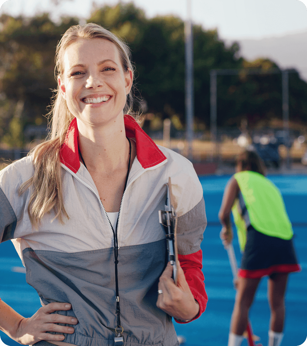 Group 1321317508 A smiling woman in a sports jacket holds a clipboard on a blue outdoor field, with another person in sports attire visible in the background.