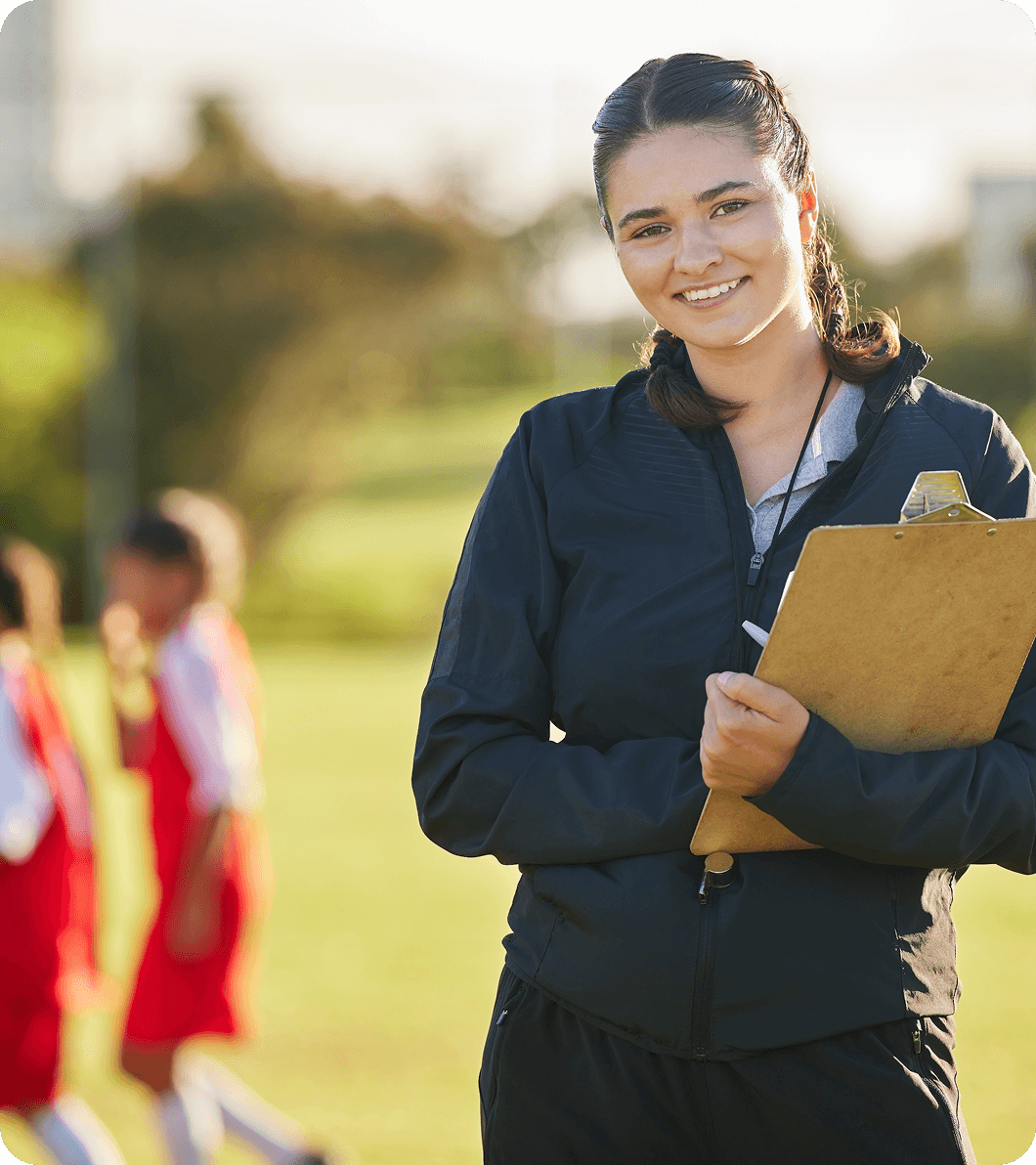 Group 1321317515 Woman holding a clipboard stands on a sports field, smiling at the camera, with children in red uniforms playing in the background.