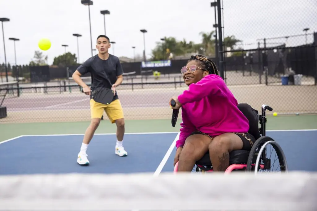 Wheelchair tennis player hitting ball, smiling opponent.